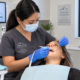 A dental professional in scrubs examines a patient’s teeth in a modern dental office. Dental tools and a screen with oral health information are visible.