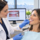 A dentist holds a tooth shade guide to a patient’s smile in a clinic, with dental images and mountains visible in the background.